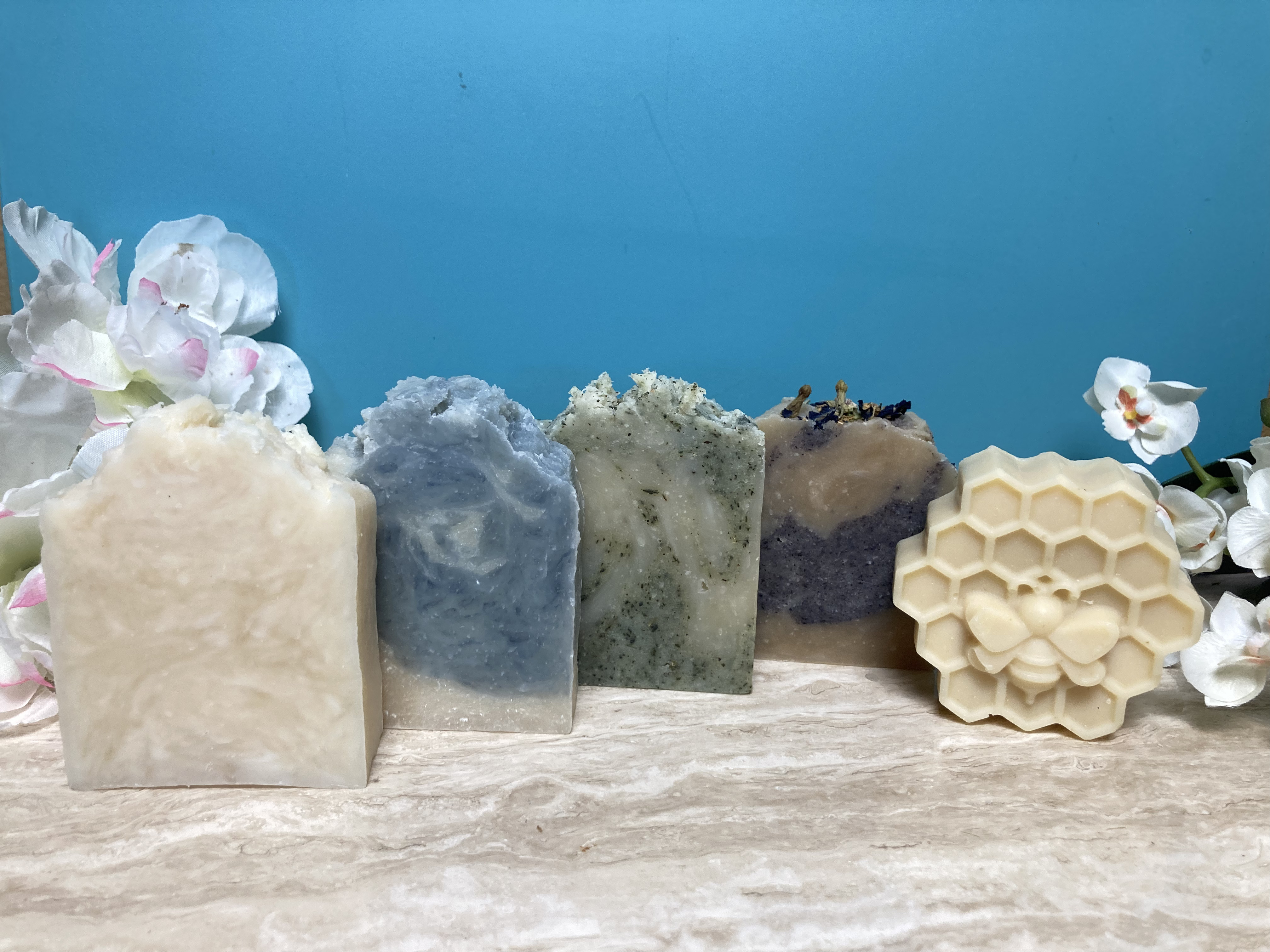 Five Sudsables signature handcrafted soaps—Mega-moisture, Blue Pine, Aloe Tallow, Lavender Swirl, and Goat Milk Oat & Honey—displayed on marble with floral accents and aqua background